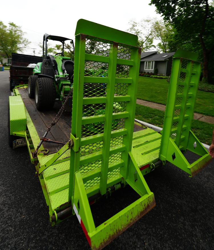 A green tree-service equipment trailer with fold-down ramps and a compact tractor at a residential job